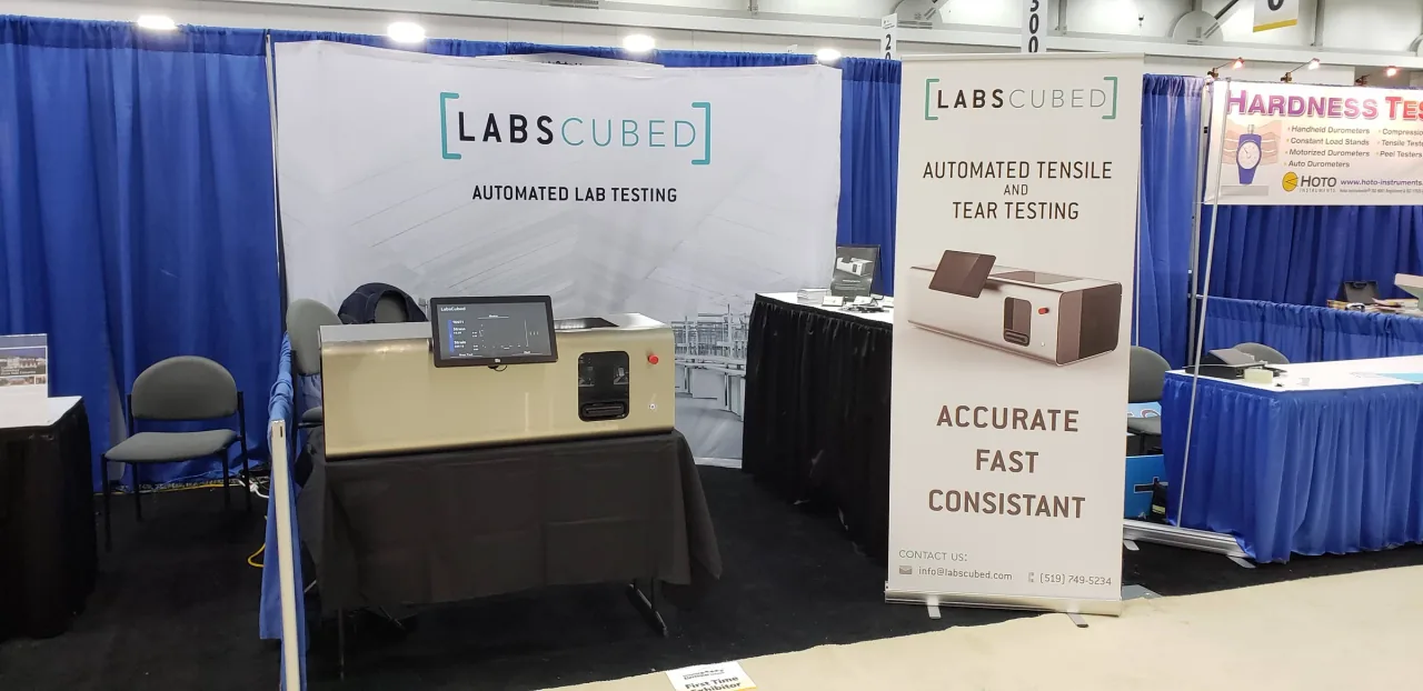 The first LabsCubed CubeOne automated rubber tensile testing machine on display at a trade show booth.
